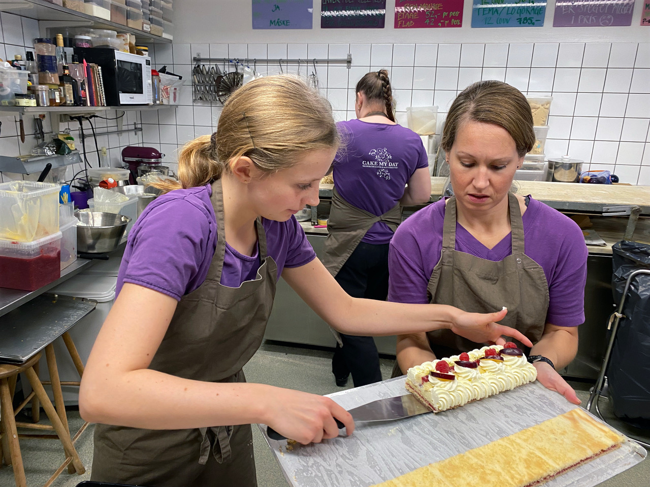 Tre ansatte i lilla t-shirts, hvor dessertdragen og logoet Cake my day er påtrykt, er i gang med at lave en konditorkage.
