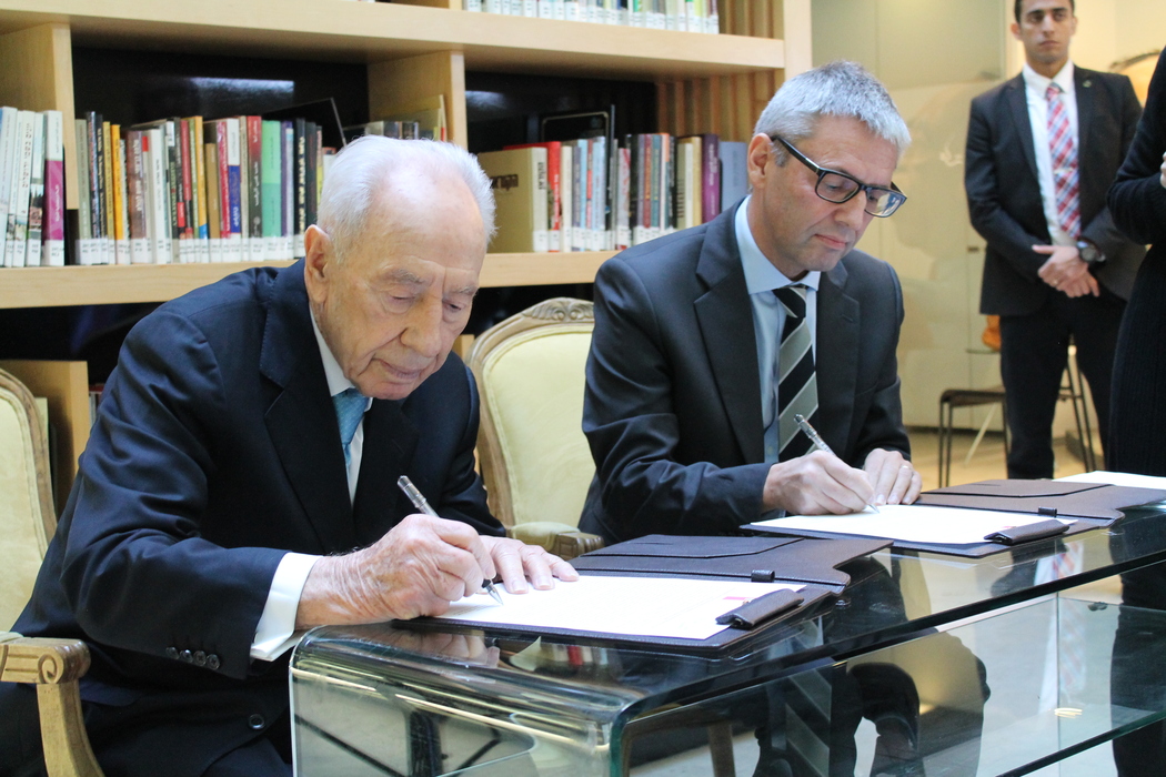 Den israelske politiker Shimon Peres sidder med Danmarks daværende ambassadør, Jesper Vahr, og underskriver en samarbejdsaftale ved et glasbord. Begge er iført jakkesæt og slips. Vahr bærer briller, og de sidder foran en bogreol. 