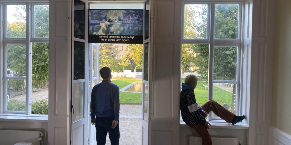 Mand står og ser op på skærm, hvor en del af forestillingen spiller. Der er udsigt til en have udenfor, og en dreng sidder i vindueskarmen