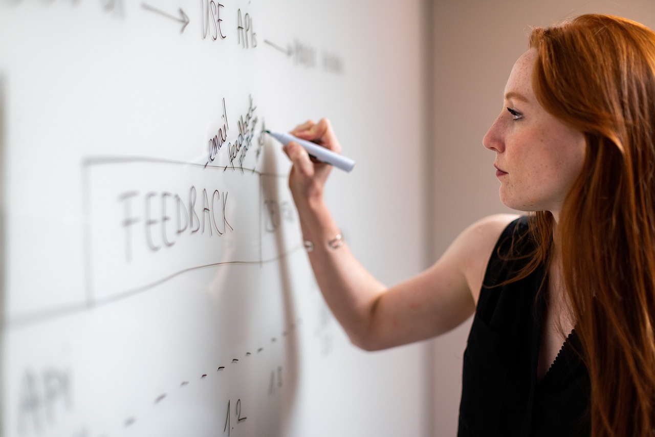 Tilpasning eller resignation? Foto af kvinde, der skriver på et whiteboard, og man ser, at der allerede er skrevet "feedback". Kvinden ser bestemt og robust ud, men ikke nødvendigvis glad og tilfreds.