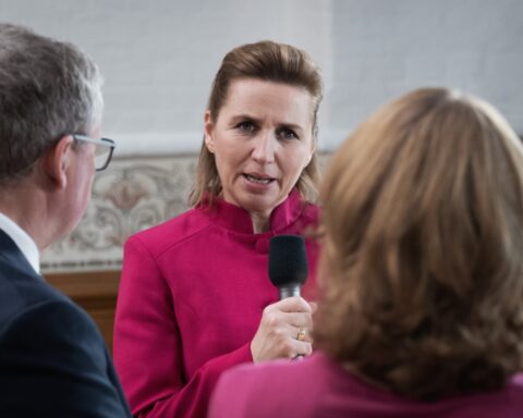 Foto af Mette Frederiksen med et skarpt udtryk, mens hun interviewes af journalister.